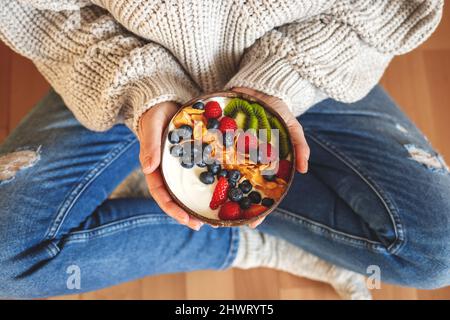 Colazione sana a casa. Donna che indossa jeans e ciotola con yogurt e fiocchi di mais ricoperti di mirtillo, lampone, kiwi a fette Foto Stock