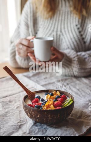 Ciotola di cocco con yogurt, fiocchi di mais e frutta fresca sul tavolo. Donna che beve caffè durante la colazione a casa Foto Stock