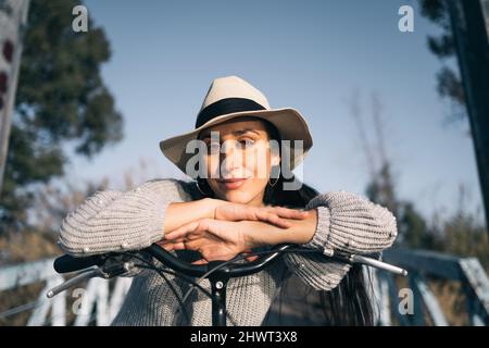 Giovane donna sorridente con cappello appoggiato sul manubrio di una bicicletta guardando la macchina fotografica Foto Stock