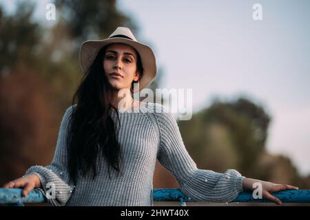 Ritratto di una donna spagnola bruna defiant guardando la macchina fotografica con un cappello su un molo vicino a un lago Foto Stock