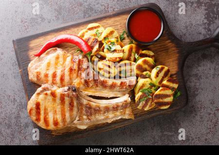 Braciole di maiale calde e succose alla griglia con strisce. Patate, peperoncino su vecchio asse di legno sul primo piano del tavolo. Vista dall'alto orizzontale Foto Stock