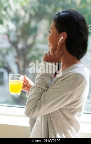 Una giovane donna positiva indossa gli auricolari quando beve una tazza di succo di frutta, guarda attraverso la finestra e chiama un amico o un collega Foto Stock