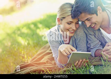 Navigazione nel cortile. Scatto di una giovane coppia felice sdraiata sull'erba e usando un tablet digitale insieme. Foto Stock