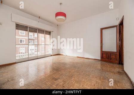 Ampio soggiorno vuoto con una grande finestra che si apre su una terrazza che si affaccia sulla strada e una porta di legno con una finestra di vetro colorato Foto Stock