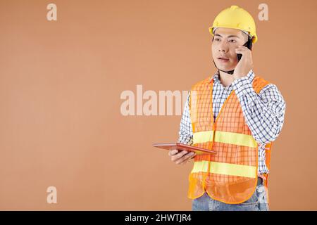 Tecnico di costruzione serio che tiene il calcolatore del tablet quando parla al telefono con il capocommessa o l'appaltatore Foto Stock