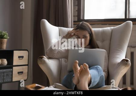 La giovane donna pensiva si siede sulla poltrona utilizzando un computer portatile Foto Stock