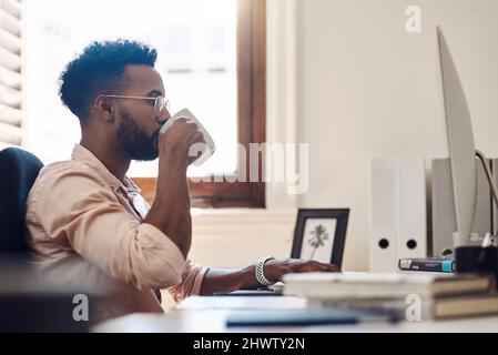 Il commercio è un gioco dove quelli con tenacia vincono sempre. Shot di un giovane uomo d'affari che beve caffè mentre lavora su un computer in un ufficio. Foto Stock