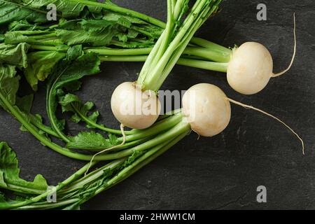 Tre bulbi di ravanello bianco con foglie verdi su ardesia nera come asse, vista dall'alto Foto Stock