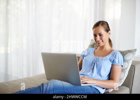 Verifica degli aggiornamenti più recenti. Scatto di una giovane donna attraente utilizzando un computer portatile in una posizione comoda sul divano. Foto Stock