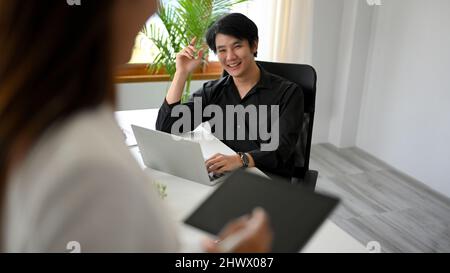 Un bel uomo d'affari siede alla scrivania dell'ufficio e sorride ad una collega femminile in ufficio. Uomo d'affari felice e allegro. Foto Stock