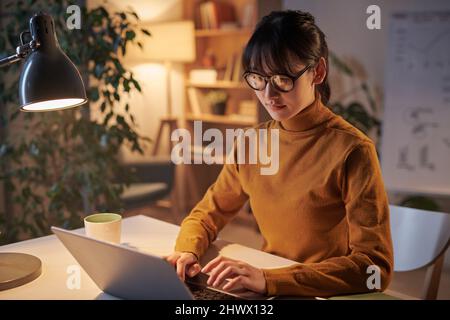 Giovane donna d'affari in occhiali concentrandosi sul suo lavoro in linea seduto sul posto di lavoro e digitando sul laptop Foto Stock