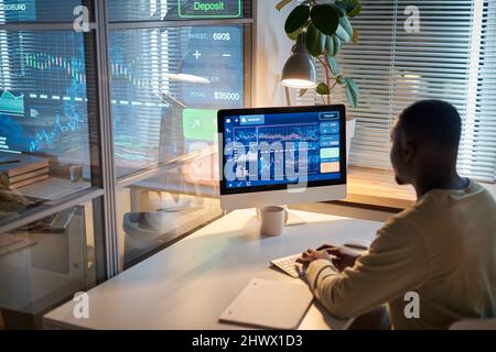 Vista posteriore del programmatore africano seduto sul posto di lavoro di fronte al computer e lavorando con i grafici sul monitor, lavora fino a tarda notte Foto Stock