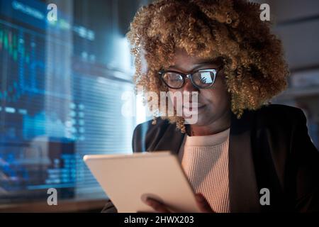 Programmatore femminile africano con capelli ricci indossando occhiali sviluppo software su tablet digitale, lavora in ufficio fino a tarda notte Foto Stock