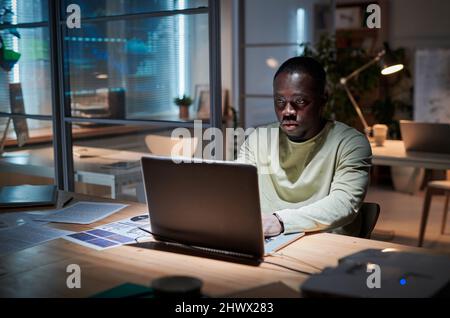 Un uomo d'affari africano che utilizza il computer portatile per prepararsi con il suo rapporto seduto al tavolo nella sala del consiglio di amministrazione e che lavora fino a tarda sera Foto Stock