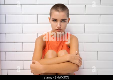 Ragazza calva in una T-shirt arancione siede sul pavimento abbracciando le ginocchia con le mani sullo sfondo di un muro di mattoni bianchi. Il concetto di cancro e solitudine nella lotta contro la malattia Foto Stock