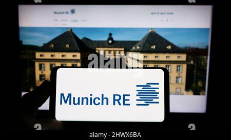 Persona che detiene il telefono cellulare con il logo della compagnia assicurativa tedesca Munich Re Group sullo schermo di fronte alla pagina web aziendale. Mettere a fuoco sul display del telefono. Foto Stock
