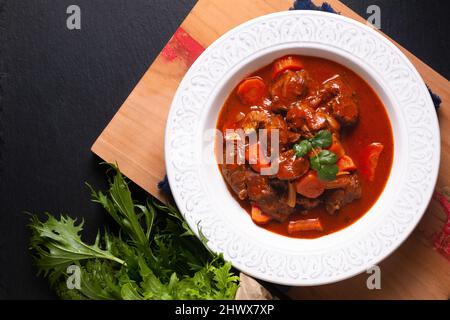 Concetto di cibo fatto in casa Francese Stew Beef Marengo in ceramica bianca piatto zuppa su asse di legno su sfondo nero ardesia pietra con spazio copia Foto Stock