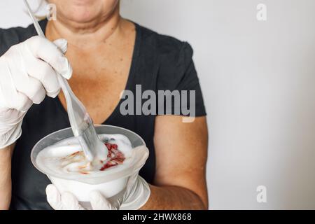 Donna anziana con ciotola trasparente piena di sostanza colorante per capelli e spazzola di plastica per tintura capelli in mani ricoperte di guanti di lattice bianco mescolando Foto Stock