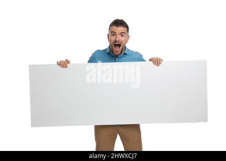 scioccato ragazzo casual con lungo cartellone bianco in mani che è sorpreso dalla pubblicità su sfondo bianco in studio Foto Stock