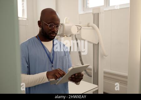 Giovane medico afroamericano in scrub medici e occhiali utilizzando il tablet mentre si guarda attraverso dati online o forma elettronica Foto Stock