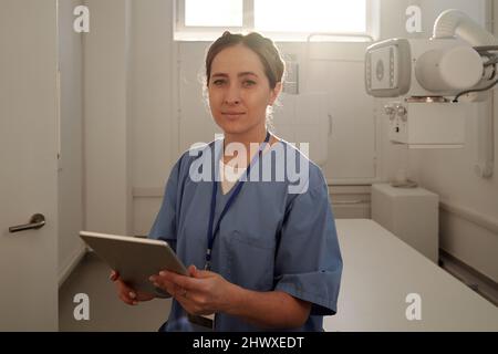Giovane radiologo femminile con tablet in piedi davanti alla telecamera contro apparecchiature a raggi X mentre lavora in ospedale Foto Stock
