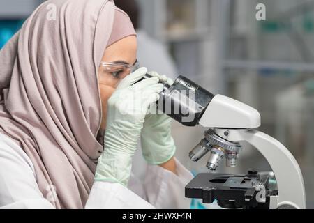 Vista laterale di giovane scienziata musulmana in hijab, guanti e occhiali protettivi studiando nuovo composto chimico in laboratorio Foto Stock