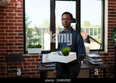 Dipendente afro-americano dall'aspetto deluso che viene licenziato lasciando il vassoio di tenuta dell'ufficio con effetti personali. Ha gettato via il lavoratore di avviamento che perde il lavoro dopo il downsizing dell'azienda. Foto Stock