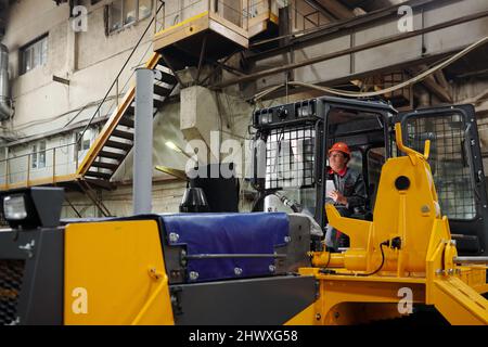 Giovane lavoratrice contemporanea in abbigliamento da lavoro e casco di sicurezza che opera enormi macchine da costruzione durante il lavoro Foto Stock