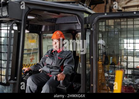 Costruttore in elmetto e abbigliamento da lavoro seduto in cabina della macchina da costruzione e guidarla mantenendo le mani sui cambi marcia Foto Stock