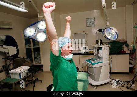 Un chirurgo affaticato stanco allunga le braccia mentre si siede su un tavolo operatorio. (MODELLO RILASCIATO) Foto Stock