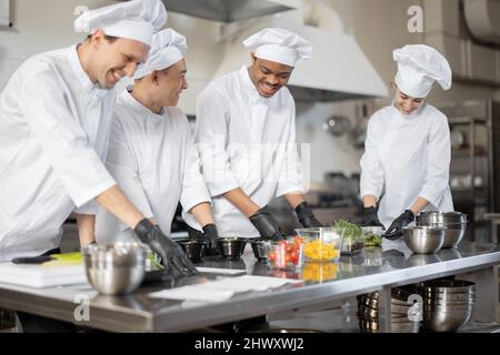 Gruppo di cuochi multirazziale che mescola gli ingredienti per portare via il cibo in cucina professionale. Concetto di cucina scura per cucinare per la consegna. Idea di lavoro di squadra in un ristorante. Persone diverse al lavoro Foto Stock