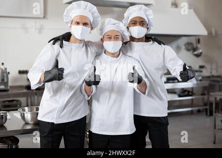 Ritratto di squadra multirazziale di tre chef in piedi insieme e mostrando OK segno. Chef ben vestiti che indossano maschere per il viso e guanti protettivi per prevenire la diffusione di infezioni durante la pandemia Foto Stock