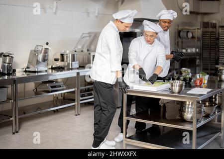 Tre cuochi multirazziali che hanno un po' di conversazione mentre cucinano in cucina professionale. Interni con attrezzature professionali e tavoli in acciaio inox. Lavoro di squadra al ristorante Foto Stock