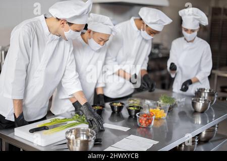 Multirazziale squadra di cuochi in maschere viso mescolando ingredienti per portare via cibo in cucina professionale. Concetto di cucina scura per cucinare per la consegna durante pandemic. Idea di lavoro di squadra in un ristorante Foto Stock