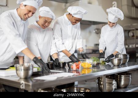 Gruppo di cuochi multirazziale che mescola gli ingredienti per portare via il cibo in cucina professionale. Concetto di cucina scura per cucinare per la consegna. Idea di lavoro di squadra in un ristorante. Persone diverse al lavoro Foto Stock