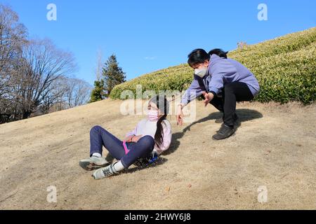 Una madre e una figlia condividono un momento di gioco nel parco, ridendo, scivolando sull'erba in una giornata di sole. Circondato da piante e alberi Foto Stock