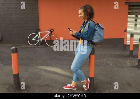 Sorridente giovane afroamericana appoggiata su un palafro usando lo smartphone Foto Stock