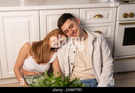 Felice giovane coppia seduta sul piano della cucina, sorridente, parlante e guardandosi, sognando, discutendo i piani felici futuri. Bella donna che mette testa su marito o spalla ragazzo. Foto di alta qualità Foto Stock