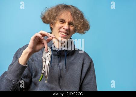 le pinze di fissaggio hanno schiacciato il dito di un adolescente con capelli ondulati vestiti con una felpa grigia . sfondo blu, spazio copia Foto Stock