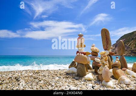 Gruppo di pietre equilibranti sulla spiaggia. Giorno di sole luminoso, atmosfera estiva, acqua di mare azzurra spruzzi su ghiaia costa, cielo blu e nuvole chiare Foto Stock