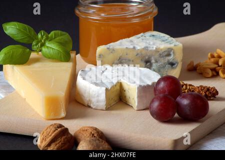 Tre tipi di formaggio, un vasetto di miele delizioso, noci, uva e basilico sono impilati su un asse di legno. La foto è stata scattata su sfondo nero. Foto Stock