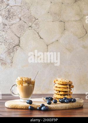 Caffè Dalgona in un bicchiere trasparente con biscotti al mirtillo e mirtilli su un asse di legno. Sfondo rustico parete. Caffè coreano molto alla moda. Foto Stock