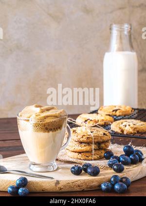 Caffè Dalgona in un bicchiere trasparente con biscotti al mirtillo e mirtilli su un asse di legno. Sfondo rustico parete. Caffè coreano molto alla moda. Foto Stock