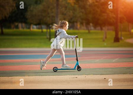 Ritratto della bambina attiva che guida scooter su strada nel parco all'aperto il giorno d'estate. Sport stagionale per bambini Foto Stock