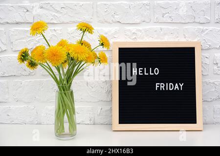 Ciao Venerdì parole su nero lettera bordo e bouquet di fiori giallo dente di leone sul tavolo contro muro di mattoni bianchi. Concept Happy Monday. Modello per Foto Stock