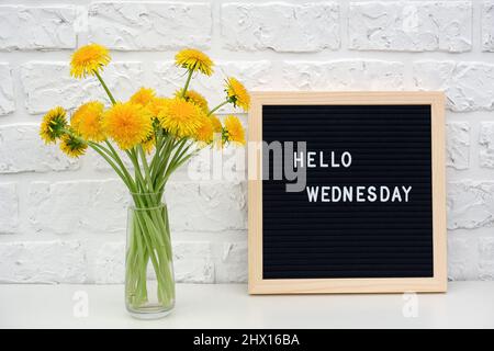 Ciao Mercoledì parole su nero lettera bordo e bouquet di fiori giallo dente di leone su tavola contro muro di mattoni bianchi. Concept Happy Monday. Modello Foto Stock