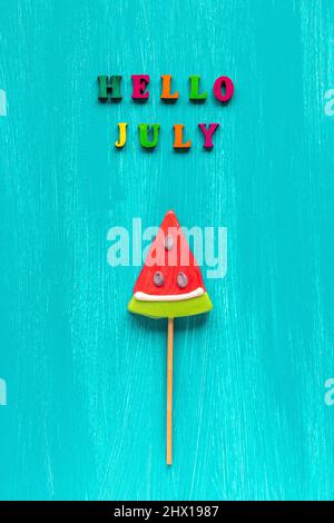 Ciao luglio testo da lettere colorate e lolllipop di anguria sul bastone su sfondo blu legno. Biglietto di auguri per il modello di visualizzazione dall'alto Concept Creative. Foto Stock