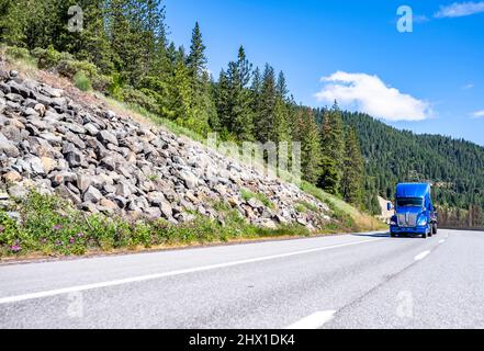 Big RIG blu industriale di grado potente trattore semi-camion con carico fissato sul semi-rimorchio a pianale che corre in salita sul moun tortuoso Foto Stock
