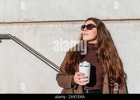 Donna in outerwear con occhiali da sole e zero spreco tazza di caffè da asporto godendo la luce del sole mentre si appoggia su parete di cemento Foto Stock