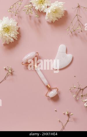 Rullo massaggio viso al quarzo rosa su sfondo rosa con fiori di gypsofila. Strumenti per massaggi con jade stone, skincar anti-invecchiamento, anti-rughe bellezza Foto Stock